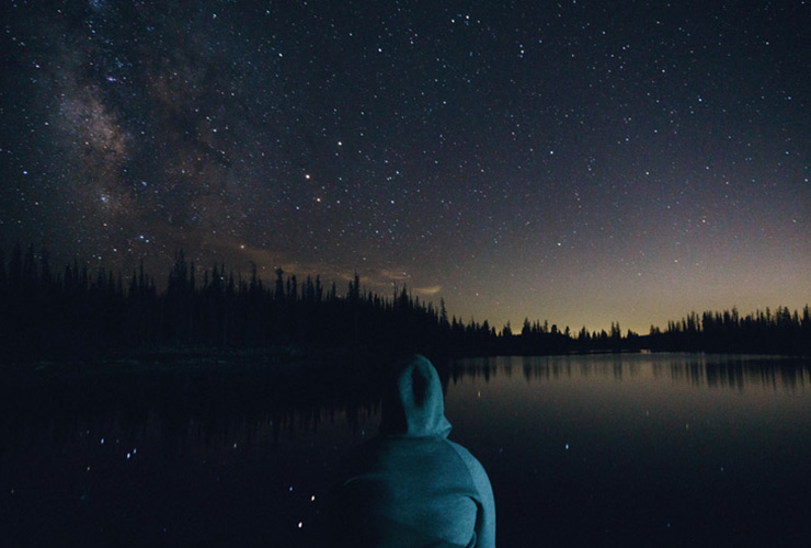 5 World Destinations for Exploring Mindfulness, Meditation and Nature by Sofia Lockart. Photograph of a man sitting by a lake looking at the starry sky by Tyson Dudley