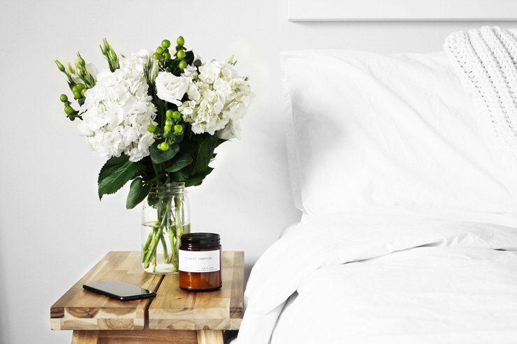 How to Create a Healthy and Safe Sleeping Sanctuary by Emma Williams. Photograph of a nightstand next to a bed, with flowers and a candle on it by Logan Nolan