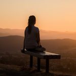 BE STILL: The Power of Stillness & Silence by Amisha Ghandiali. Photograph of a woman sitting on a mountain top at sunset by Sage Friedman
